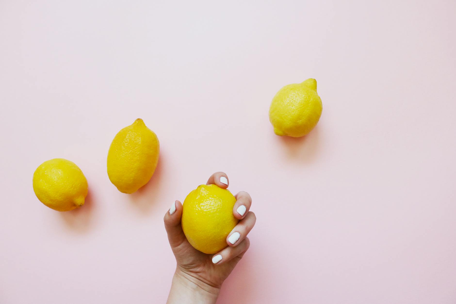 A hand holding a lemon on a soft pink background, representing natural pleasure and vitality